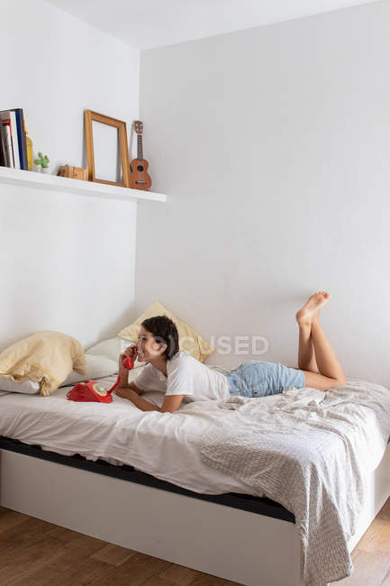 Piacevole donna sorridente e rispondente telefonata da vintage telefono rosso mentre sdraiato sul letto in camera da letto accogliente — Foto stock