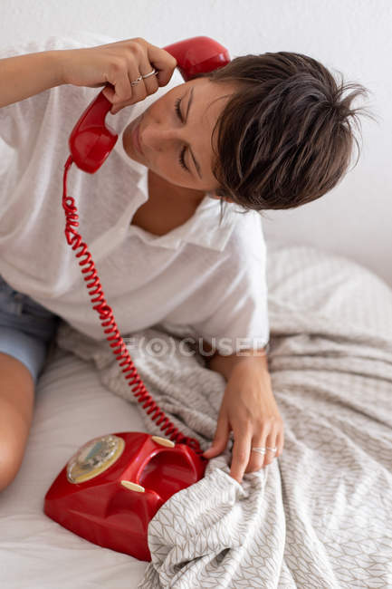 Woman answering phone call from vintage red telephone while sitting on bed in cozy bedroom — Stock Photo