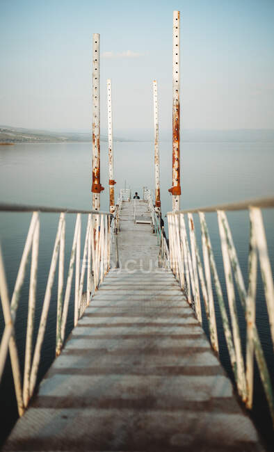 Narrow pier with weathered railings and distant traveler located near tranquil sea water against cloudless sky — Stock Photo