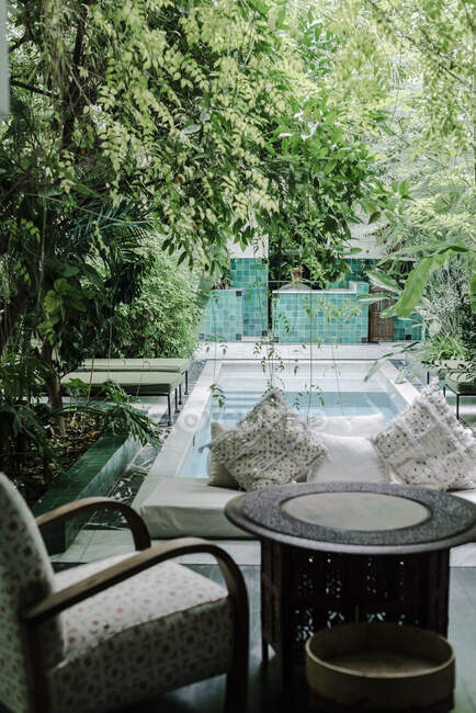 Banco suave con almohadas situado cerca de la piscina limpia en el jardín verde del hotel en Marrakech, Marruecos - foto de stock