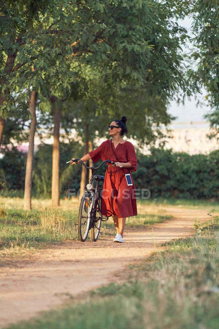 Woman in dress pushing bike and smiling while walking along path on sunny day in park — Stock Photo