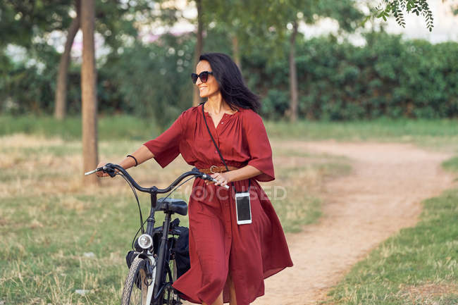 Full length woman in dress pushing bike and smiling while walking along path on sunny day in park — Stock Photo