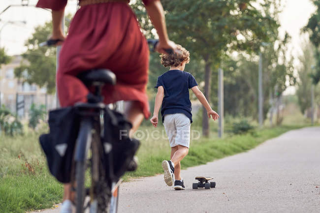 Menino andar de skate na frente da mulher alegre na bicicleta ao longo do caminho asfalto enquanto passar o tempo no parque juntos — Fotografia de Stock