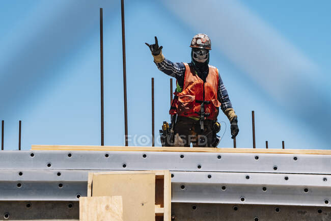 Человек в маске и в каске в ярком жилете с инструментами, делающими жест скалы, стоя на вершине развивающегося здания — стоковое фото