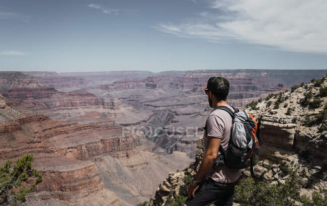 Vista lateral del hombre con mochila contemplando y de pie en la cima de la montaña en el caluroso día de verano en EE.UU. - foto de stock