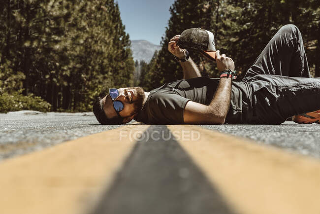 Side view of positive bearded man in sunglasses smiling and lying on back on concrete lane surrounded with leafy trees in USA — Stock Photo