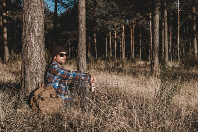 Vista lateral del hombre con sombrero y gafas de sol apoyado en el árbol de hoja perenne y sentado en la hierba por la mochila en el día soleado - foto de stock