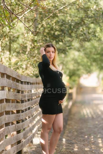Mulher grávida olhando para a câmera e tocando barriga enquanto está de pé na estrada perto do jardim em dia ensolarado — Fotografia de Stock