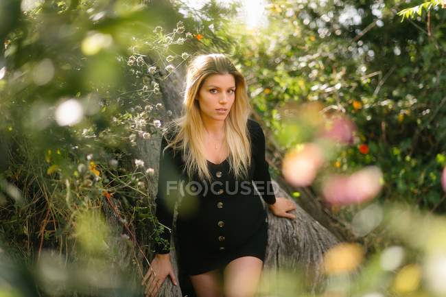Спокойная привлекательная беременная женщина в элегантном черном платье смотрит в камеру, сидя среди цветов в саду — стоковое фото