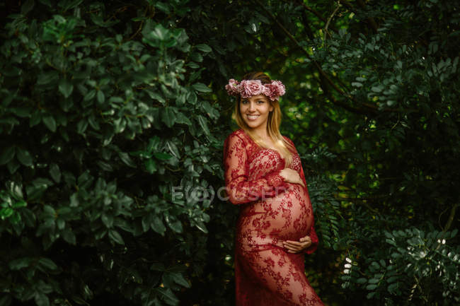 Mulher grávida com os olhos fechados tocando barriga enquanto está em pé no jardim no dia ensolarado — Fotografia de Stock