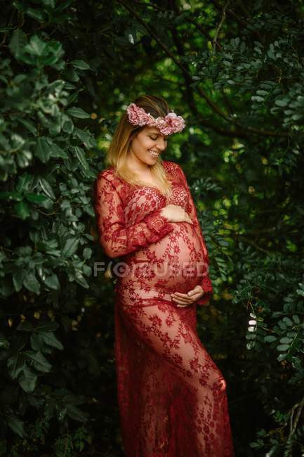 Mulher grávida com os olhos fechados tocando barriga enquanto está em pé no jardim no dia ensolarado — Fotografia de Stock