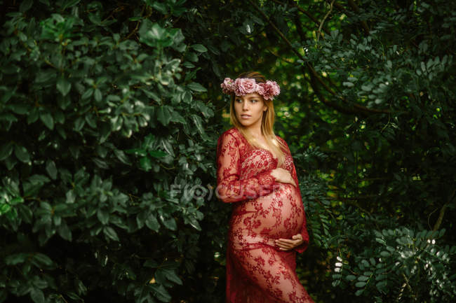Mujer embarazada con los ojos cerrados tocando el vientre mientras está de pie en el jardín en el día soleado - foto de stock