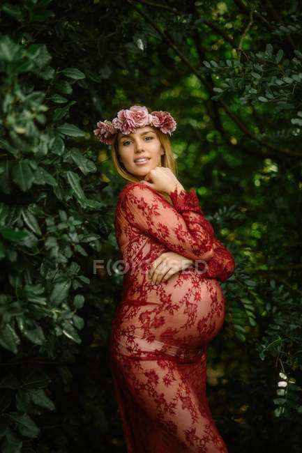 Беременная женщина с закрытыми глазами касается живота, стоя в саду в солнечный день — стоковое фото