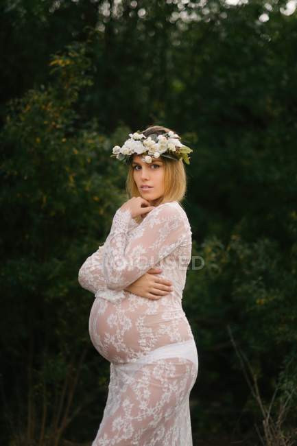 Pregnant woman with closed eyes in white lace dress touching chin while standing in forest — Stock Photo