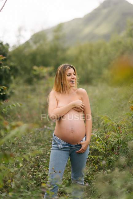 Pregnant topless woman looking away and covering breast while standing in meadow — Stock Photo