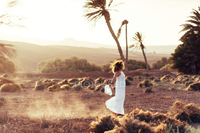Vista lateral da mulher de vestido branco andando em campo com grama seca em Fuerteventura, Las Palmas, Espanha — Fotografia de Stock