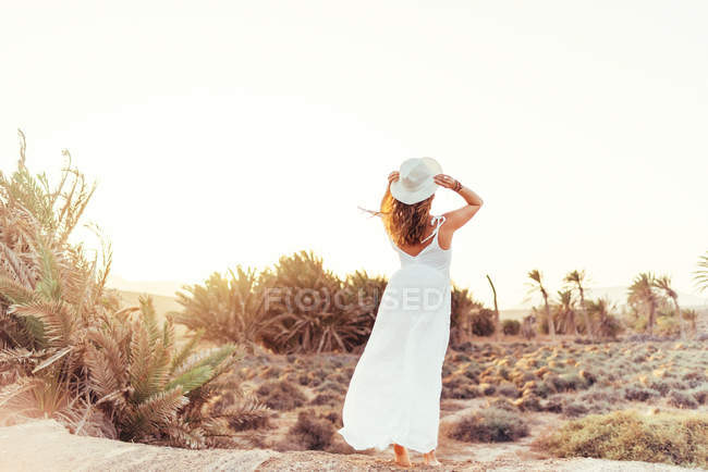 Woman in white dress touching hat in dry field in sunlight — Stock Photo