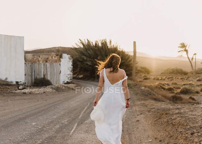 Mulher de vestido branco na estrada rural com campo seco à luz do sol — Fotografia de Stock