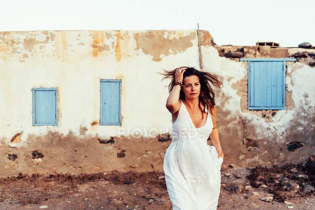 Incredibile donna in abito bianco toccare i capelli dalla vecchia casa squallida con le finestre blu chiuse a Fuerteventura, Spagna — Foto stock