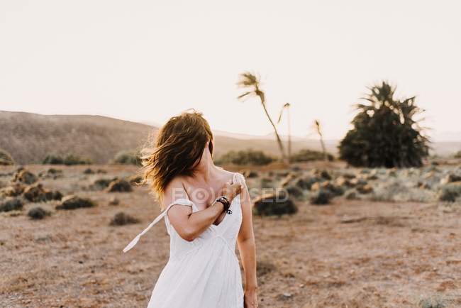 Femme en robe blanche avec des cheveux salissants debout dans un champ sec au soleil — Photo de stock