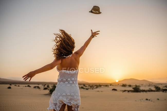 Visão traseira da mulher magra levantando as mãos e jogando chapéu ao pôr do sol no vento em Fuerteventura, Las Palmas, Espanha — Fotografia de Stock