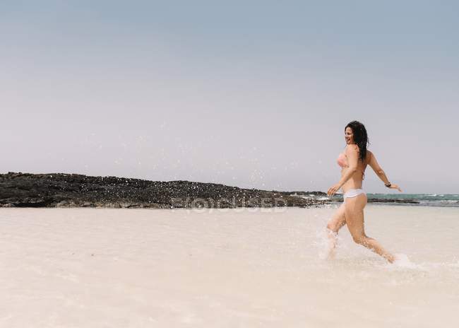 Vista lateral de una mujer alegre corriendo en aguas cristalinas y salpicando en la playa en Fuerteventura, Las Palmas, España - foto de stock