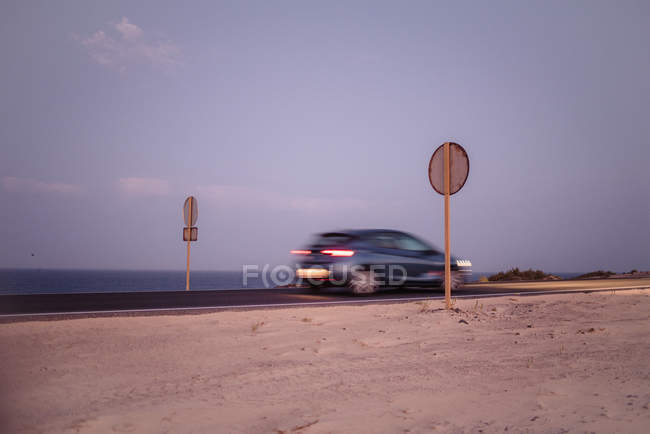 Coche azul que circula rápido por carretera con señales redondas sobre pilares de madera a lo largo del océano en Fuerteventura, Las Palmas, España - foto de stock