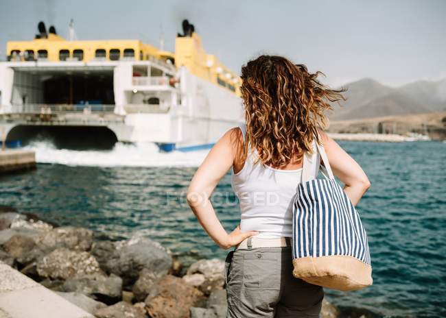 Visão traseira da mulher ativa com as mãos de lado olhando para o barco grande em ondas turquesa no cais em Fuerteventura, Las Palmas, Espanha — Fotografia de Stock