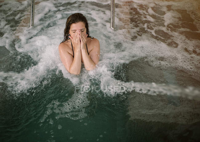 Сверху женщина с закрытыми глазами в бикини закрывает лицо от воды во время купания в бассейне с струями и пузырьками — стоковое фото