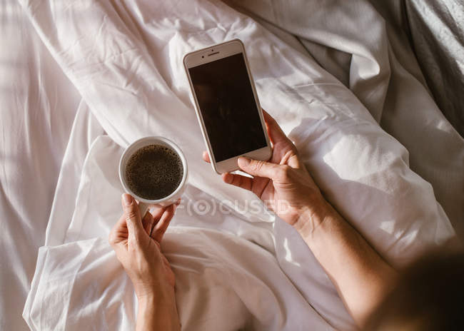 Da sopra le mani della donna utilizzando smartphone a letto e tenendo in mano una tazza di caffè al mattino — Foto stock