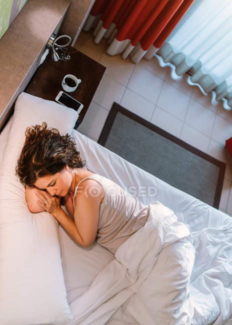 De cima calma fêmea dormindo na cama branca perto da mesa de cabeceira com xícara redonda de café e smartphone de manhã — Fotografia de Stock
