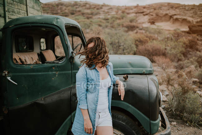 Young woman in shorts and denim shirt standing by green truck and looking away in nature — Stock Photo