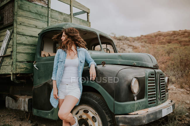 Mujer joven en pantalones cortos y camisa de mezclilla de pie junto al camión verde y mirando hacia otro lado en la naturaleza - foto de stock