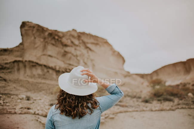 Обратный вид неузнаваемой женщины в повседневной одежде, держащей шляпу во время прогулки по песчаным дюнам пустыни — стоковое фото