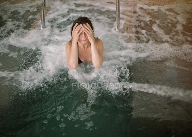 From above adult female with closed eyes in bikini covering face from water while swimming in pool with jets and bubbles — Stock Photo