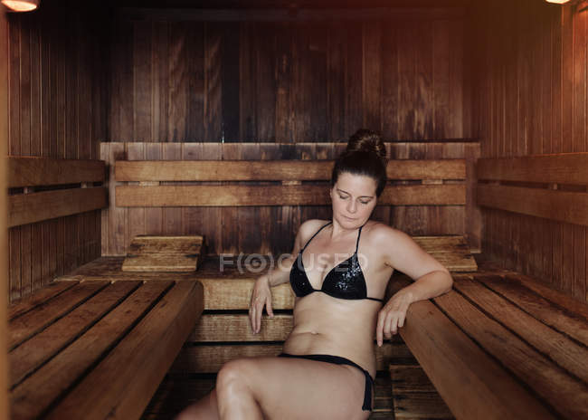 Donna in bikini nero seduta con gli occhi chiusi su un asciugamano in bagno turco appoggiata su una panchina di legno e godendo di calore — Foto stock