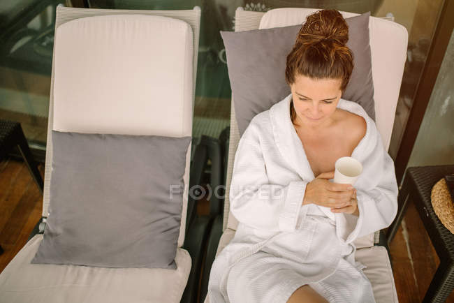 D'en haut femme en peignoir blanc boire de l'eau tout en étant couché sur la chaise longue après la procédure de spa et souriant le jour — Photo de stock