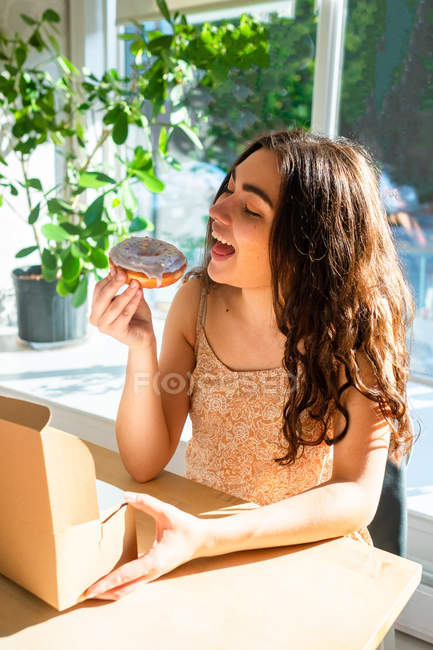 Zufriedene Frau in Sonnenhut beißt mit geschlossenen Augen in glasiertes Gebäck, während sie am Fenster am Tisch sitzt — Stockfoto