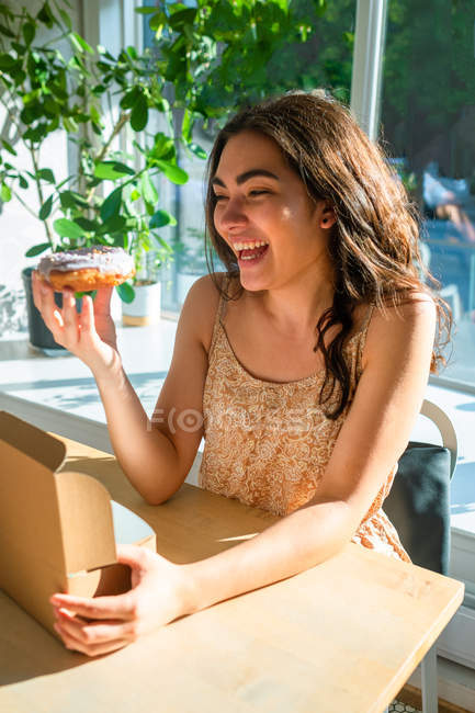 Content woman in sundress holding glazed pastry while sitting at table by window — Stock Photo