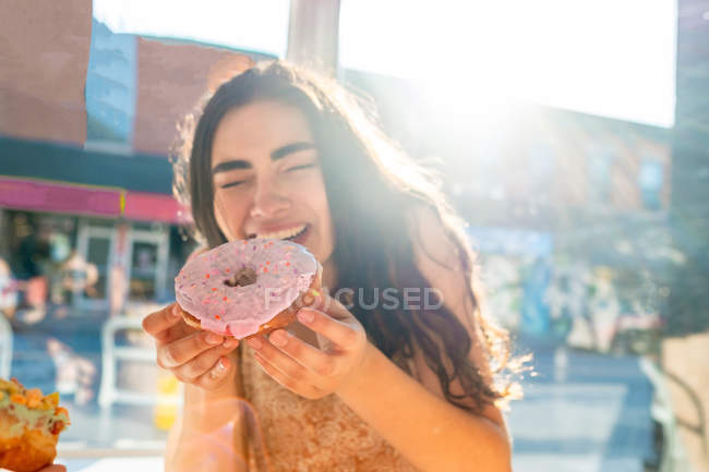 Content woman in sundress biting glazed pastry while sitting at table by window in sunlight — Stock Photo