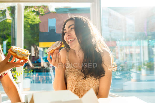 Happy women eating glazed desserts while sitting at table by window in cafeteria — Stock Photo