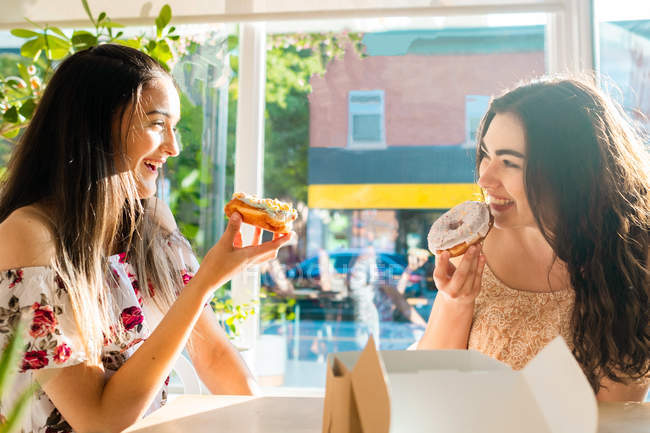Mujeres felices en vestidos de sol comiendo postres glaseados mientras se sientan en la mesa por la ventana en la cafetería - foto de stock
