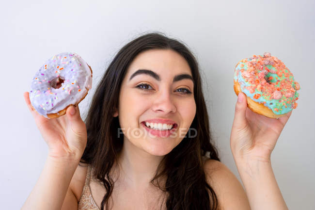 Carefree woman having fun and playing with glazed donuts with sprinkles while standing by white wall — Stock Photo