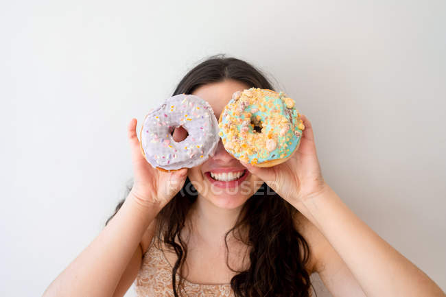Carefree woman having fun and playing with glazed donuts with sprinkles while standing by white wall — Stock Photo