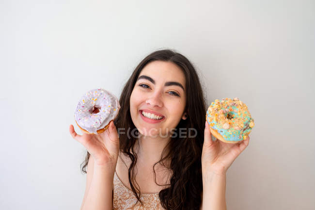 Carefree woman having fun and playing with glazed desserts with sprinkles while standing by white wall — Stock Photo