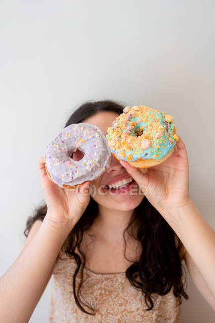 Безтурботна жінка розважається і грає з глазурованими пончиками зі зморшками, стоячи біля білої стіни — стокове фото