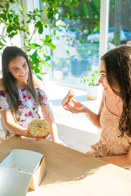 Счастливые женщины в солнечных платьях едят глазированные десерты, сидя за столом за окном в кафетерии — стоковое фото