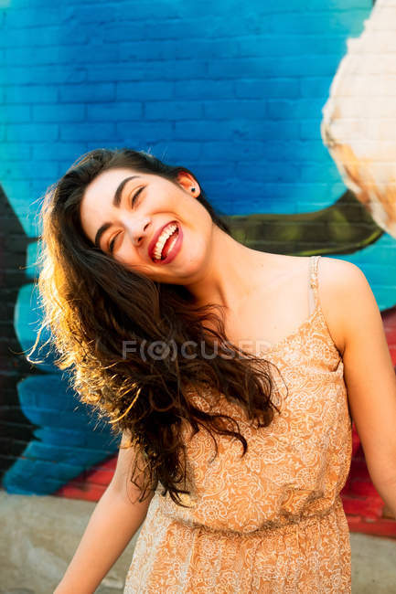 Donna giocosa in abito divertendosi e inclinando la testa sul lato mentre in piedi vicino al muro di mattoni urbani — Foto stock