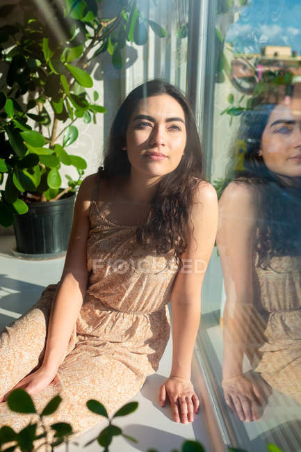 Dreamy woman in sundress contemplating while sitting on window sill with greenery — Stock Photo
