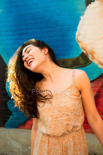 Verspielte Frau im Kleid, die Spaß hat und den Kopf auf die Seite neigt, während sie an einer städtischen Ziegelmauer mit Graffiti steht — Stockfoto